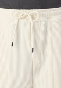Cremefarvede ribbede bukser med elastisk talje, der har en snorlukning med sorte spidser. Blød tekstur med sidelommer.