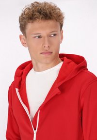 Rode hoodie met ritssluiting en een zachte, gestructureerde binnenkant, voorzien van een witte rits en een capuchon. Draag het over een wit shirt, wat de halslijn benadrukt.