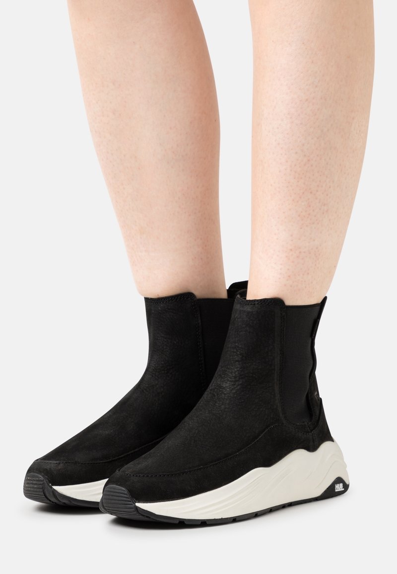 Personne portant des bottes noires mi-mollet avec des semelles blanches épaisses et des panneaux élastiques, debout sur un fond blanc.