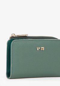 Portafoglio in pelle verde con finitura testurizzata, caratterizzato da un logo tono oro, chiusura con zip e forma rettangolare compatta.