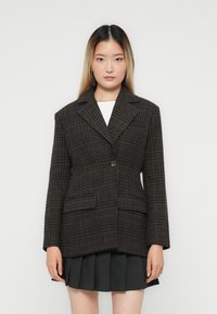 Jonge vrouw in een donker geruite blazer over een witte top en een zwarte geplooide rok, staand tegen een effen lichte achtergrond.