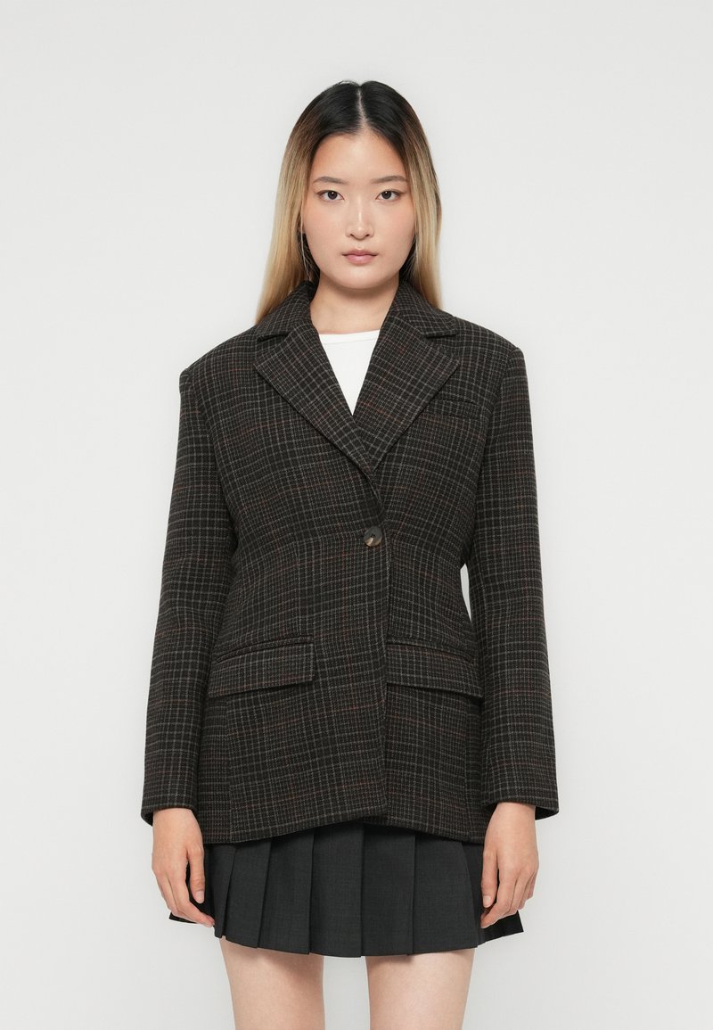 Jonge vrouw in een donker geruite blazer over een witte top en een zwarte geplooide rok, staand tegen een effen lichte achtergrond.
