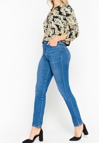 Svart blus med paisleymönster som matchas med blå tajta jeans; blusen har rund halsringning och trekvartsärmar, jeansen är formtillpassade.