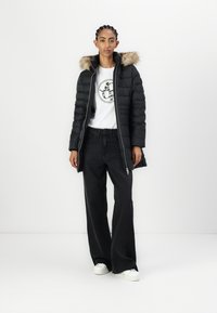 Crna puf jakna s kapuljačom od krzna, uparena s bijelom majicom koja ima okrugli grafički uzorak i crnim širokim trapericama. Bijele tenisice upotpunjuju izgled.