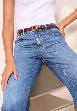 Henkilö, jolla on vaaleansiniset korkeavyötäröiset farkut, ruskea vyö ja valkoinen hihaton paita, istuen ja käsivarret lantiolla.