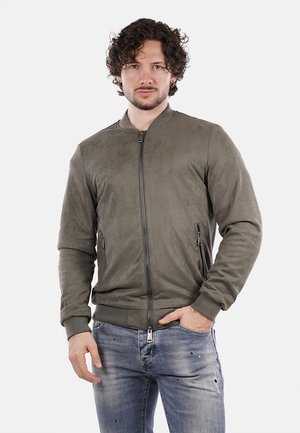 Giacca bomber in suede verde oliva con zip frontale, polsini a coste e due tasche laterali con zip, indossata sopra jeans blu chiari strappati.