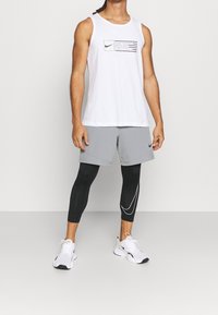 Biała, bezrękawna koszulka Nike z graficznym nadrukiem; szare szorty; czarne legginsy z białymi akcentami; białe buty Nike z czarnymi detalami.