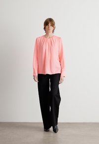 Rosa Bluse mit gerafftem Ausschnitt, ergänzt durch schwarze, weite Hosen mit dezentem Muster. Das Model steht auf einem neutralen Boden.
