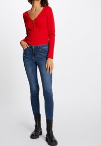 Rode geribbelde lange mouwtop met een V-hals en ritssluiting, gedragen met strak blauwe jeans en zwarte enkellaarsjes.