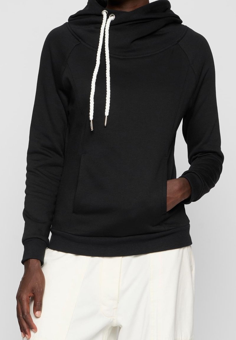Personne portant un sweat à capuche noir avec des cordons blancs et un pantalon blanc, une main dans la poche, fond gris neutre.