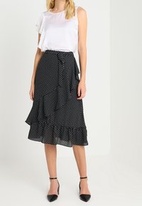 Černá sukně s puntíky a volánovým lemem, vyrobená z lehké látky; doplněná o jednoduchou bílou blůzu a černé podpatky.