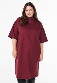 Vinröd oversized klänning av mjukt tyg, med hög krage och dragkedjeförslutning, korta ärmar och sidospjäll för rörelsefrihet.