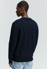 Een jonge man in een marineblauwe trui en lichte jeans is van achteren te zien tegen een lichtgrijze achtergrond.