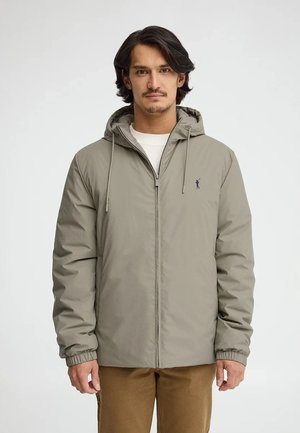 Mann mit mittellangen dunklen Haaren, der eine hellgraue Kapuzenjacke mit Reißverschluss und braune Hose trägt, vor einem einfarbigen hellen Hintergrund.