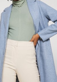 Cappotto di lana azzurro chiaro sopra un dolcevita verde a costine, abbinato a pantaloni aderenti crema. Il cappotto presenta un colletto a punta e tasche laterali.