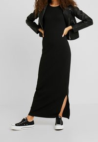 Kvinna som bär en svart läderjacka, en ärmlös svart maxiklänning med sidoklyvning, och svarta och vita plattformssneakers, stående med händerna på höfterna.