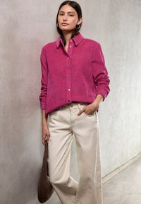 Camicia in corduroy fucsia con colletto, bottoni sul davanti e maniche lunghe, abbinata a pantaloni beige a gamba larga e una borsa marrone.