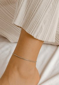 Una pulsera de tobillo de plata reposa sobre piel desnuda, combinada con una tela de ribete ligero. El primer plano enfatiza la textura suave y el diseño delicado.