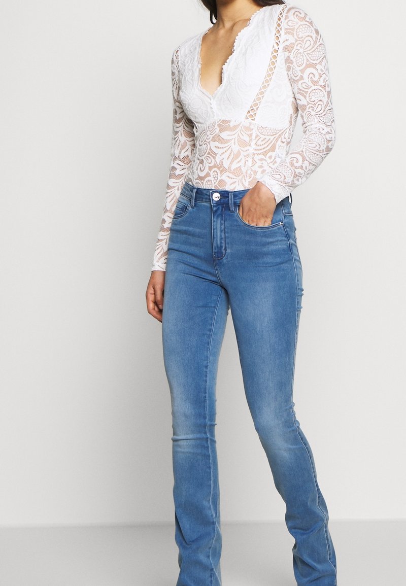 Femme portant un haut à manches longues en dentelle blanche avec un col en V et un jean bleu ajusté, debout avec une main dans la poche.