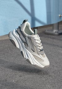 Bijela i siva chunky tenisica s mrežastim detaljima i debelim đonom, označena sa "Steve Madden", lebdi iznad asfaltne površine pored plave teksturirane zidne pozadine.