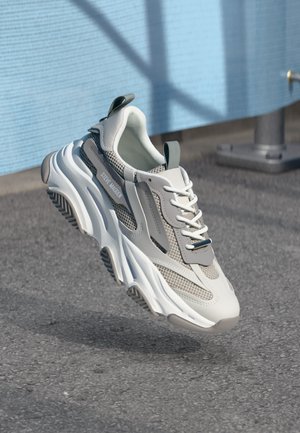 White and gray chunky sneaker with mesh details and thick sole, labeled "Steve Madden," floating above asphalt near a blue textured wall.