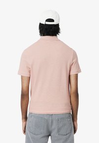 Ljust rosa polo-tröja i mjukt tyg med korta ärmar och krage. Bärs med grå jeans med bakfickor. Bakvy visas.