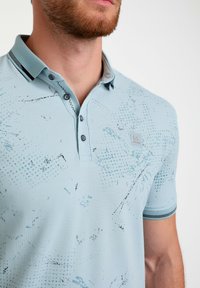 Gaiši zila polo krekla ar teksturētu rakstu, trīs pogām, kontrastējošu svītrainu apkakli un mazu logotipu uz krūtīm.