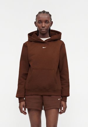 Vrouw met gevlochten haar die een bruine Nike-hoodie en bijpassende short draagt, staand tegen een eenvoudige lichte achtergrond.