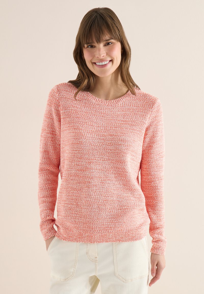 Femme souriante aux cheveux bruns portant un pull en tricot corail et un pantalon crème, debout avec une main dans la poche devant un fond beige.