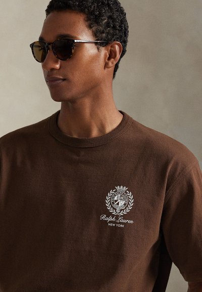 Καφέ φούτερ με στρογγυλή λαιμόκοψη, ριμπ μανσέτες και λευκό λογότυπο με στεφάνι και κείμενο. Το μοντέλο φοράει σκούρα, στρογγυλά γυαλιά ηλίου.