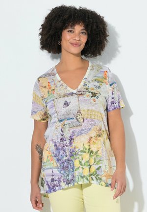 Frau mit lockigem Haar, die ein buntes V-Ausschnitt-Shirt mit Blumen- und Schmetterlingsmuster und blassgelbe Hosen trägt, steht vor einer weißen Wand.