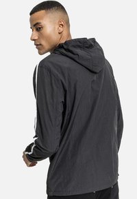 Chaqueta con capucha negra, hecha de material ligero, con mangas largas que presentan acentos blancos y un acabado texturizado. Diseño clásico, silueta simple.
