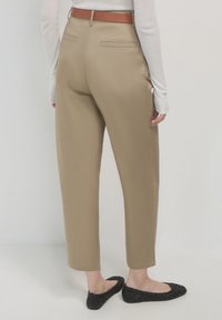 Pantalons beiges avec une texture lisse, design de jambe fuselée et poches arrière. Une ceinture marron complète la tenue. Des ballerines noires sont portées.