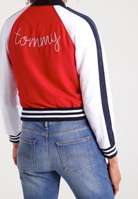Rød og hvid bomberjakke med marineblå detaljer, prydet med "tommy" broderet i skrifttype på ryggen. Ribbet kant og ærmer. Sammen med blå jeans.
