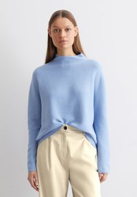 Hellblauer Pullover mit hohem Kragen, abgesenkten Schultern und abgerundetem Saum, kombiniert mit beigen Hosen. Weiche Textur, minimalistisches Design.