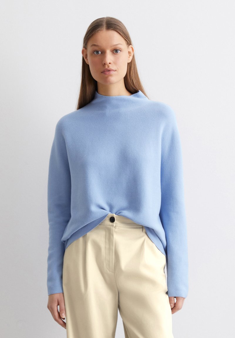 Hellblauer Pullover mit hohem Kragen, abgesenkten Schultern und abgerundetem Saum, kombiniert mit beigen Hosen. Weiche Textur, minimalistisches Design.