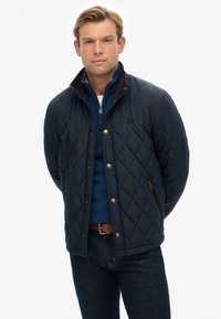 Giubbotto trapuntato blu navy con motivo a rombi, presenta un colletto in velluto a coste marrone, bottoni frontali e cerniere laterali, abbinato a un maglione blu con zip.