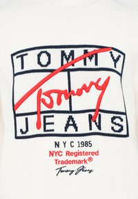 Weißer Strickpullover mit einem großen roten "Tommy Jeans"-Logo mit marineblauen Akzenten, umgeben von einem rechteckigen Rand und dem Jahr "NYC 1985".