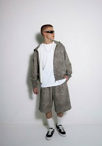 Szary zestaw z nadrukiem w postaci bluzy z kapturem i szortów z wzorem paisley, połączony z białą koszulką i czarnymi sneakersami. Uzupełniony okularkami przeciwsłonecznymi.