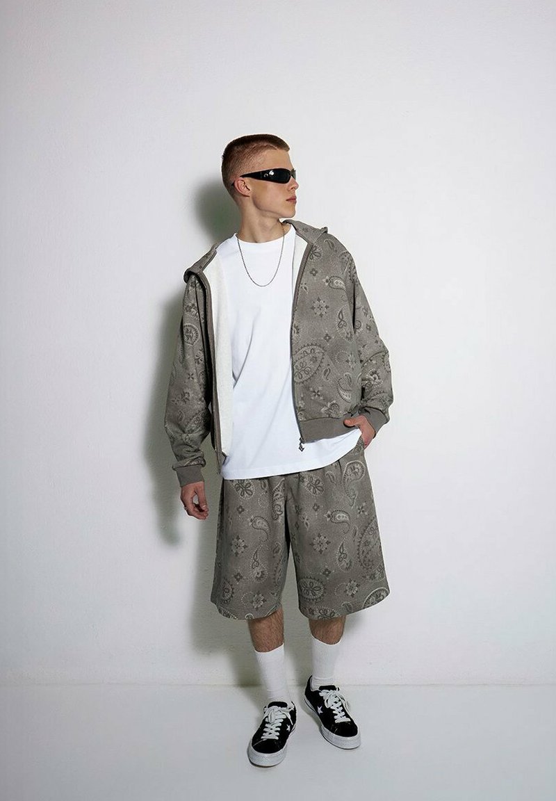 Szary zestaw z nadrukiem w postaci bluzy z kapturem i szortów z wzorem paisley, połączony z białą koszulką i czarnymi sneakersami. Uzupełniony okularkami przeciwsłonecznymi.