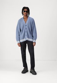 Överdimensionerad ljusblå ribbad cardigan med v-ringning och knappar, accentuerad med vita ränder på manchetterna. Matchad med svarta byxor och sneakers.