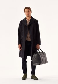 Black trench coat, light beige polo, dark jeans, green high-top sneakers, carrying a black leather bag with dual handles and zipper detail.
