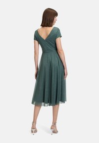 Zielona błyszcząca sukienka z krótkimi rękawami, dekoltem w kształcie litery V i rozkloszowaną spódnicą. Uzupełniona przezroczystą narzutą i noszona z srebrnymi szpilkami.