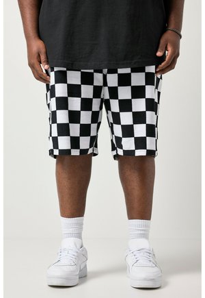 Person iført sort-hvide ternede shorts, hvide sokker, hvide sneakers og en sort skjorte med hænderne ned langs siderne.