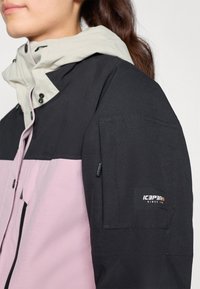 Schwarze und rosa Kapuzenjacke mit einer hellgrauen Kapuze, Fronttaschen und einem seitlichen Armaufnäher mit Branding. Glatte Textur und modernes Design.