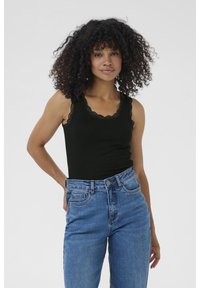 Junge Frau mit lockigem Haar trägt ein schwarzes Top und hochgeschnittene blaue Jeans, steht selbstbewusst und lächelt vor einem hellen Hintergrund.