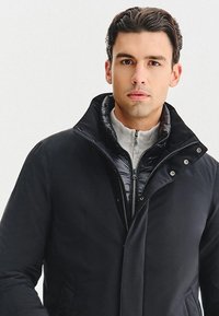 Czarna ocieplana kurtka z wysokim kołnierzem, z zapięciem na zamek błyskawiczny i zatrzaskami, warstwowa z szarym swetrem. Gładka tekstura.