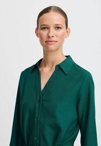 Grüne Knopf-leiste Hemd mit V-Ausschnitt und Kragen; glattes, leichtes Material mit leicht glänzender Textur; hat lange Ärmel.