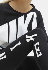 Sportklädeset som består av en svart sweatshirt med en stor vit Nike-logga och svarta leggings med vit text. Mjukt tyg och åtsittande design.