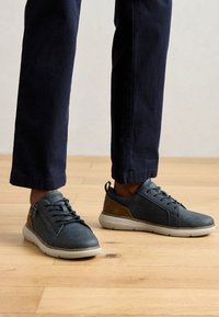 Baskets en cuir bleu avec des lacets gris et un accent marron au talon. Surface texturée, semelle en caoutchouc, et un petit détail de fermeture éclair sur le côté.